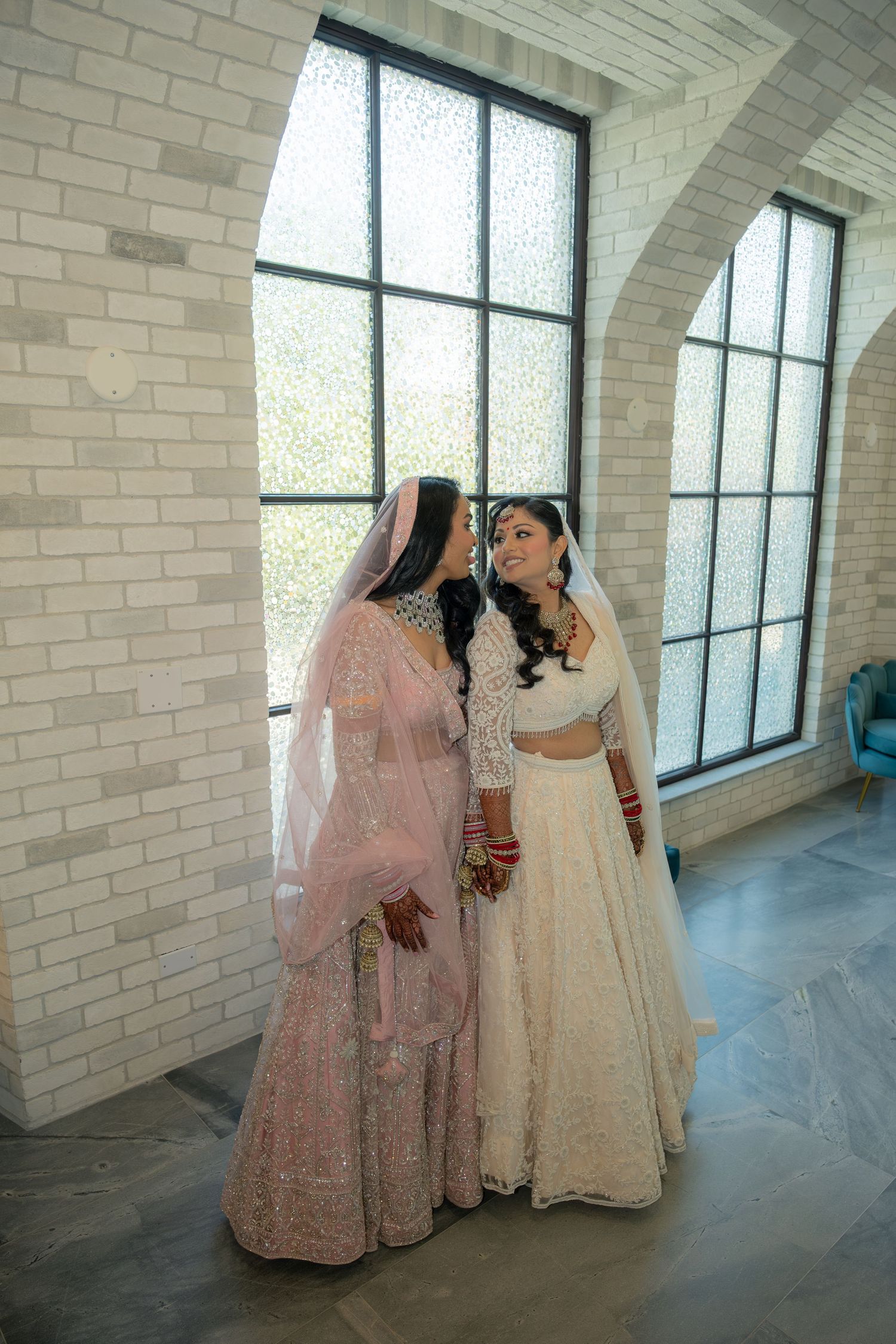 Two Indian brides in traditional white and pink lehengas share a tender moment by arched windows in a brick venue.