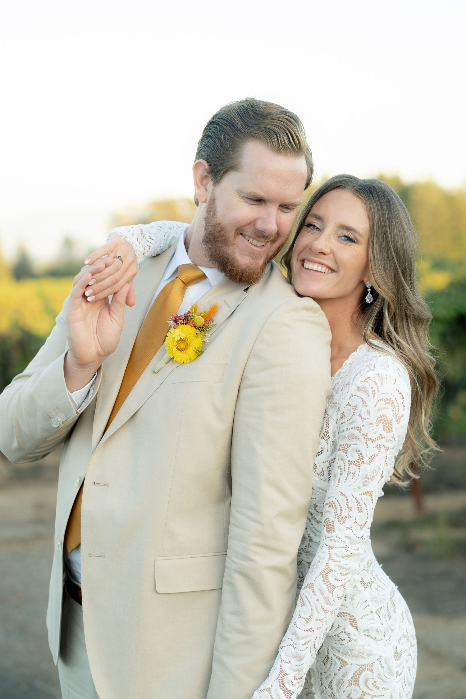 Smiling bride in lace gown embraces groom in tan suit with yellow floral boutonniere at sunset.