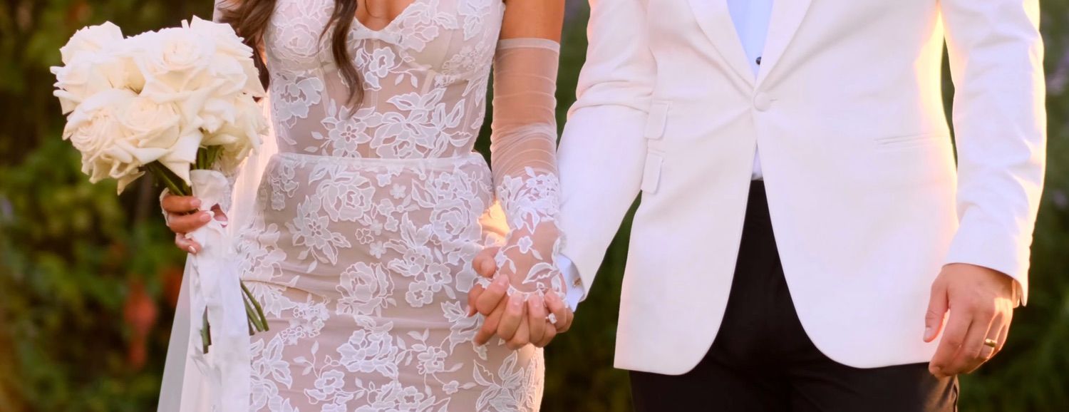 Bride in lace wedding gown and groom in white suit holding hands with a bridal bouquet of white flowers at outdoor wedding.