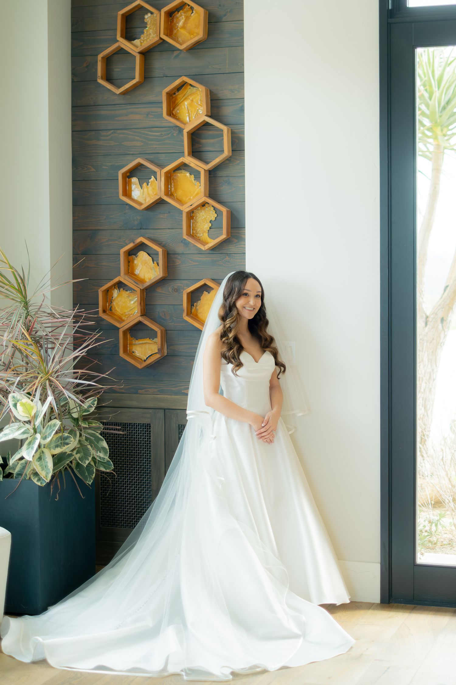 Beautiful bride in a flowing white satin ball gown with veil posing near honeycomb wooden wall decor.