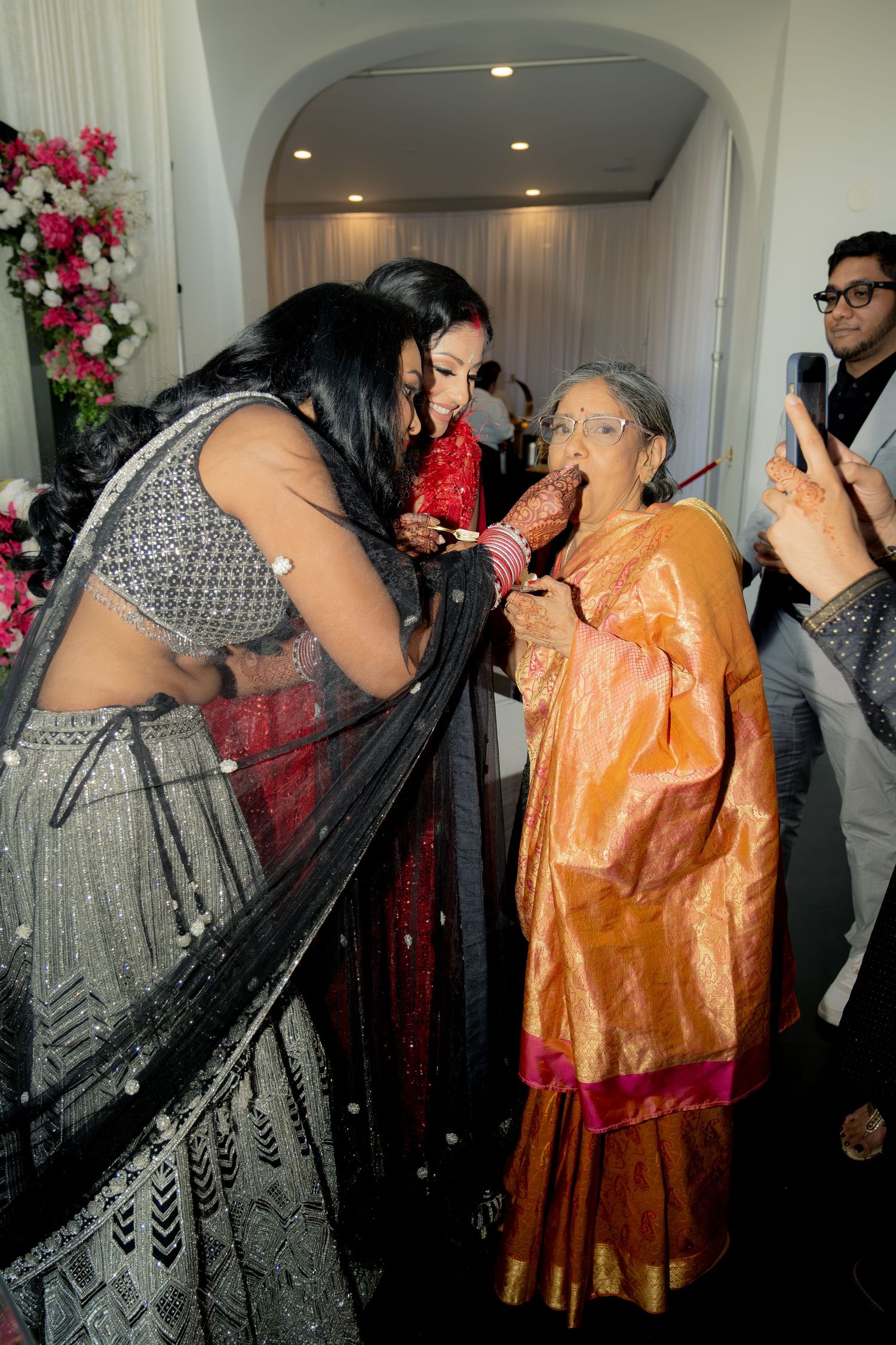 Elderly woman in orange saree blesses bride at Indian wedding ceremony while guests capture the moment.