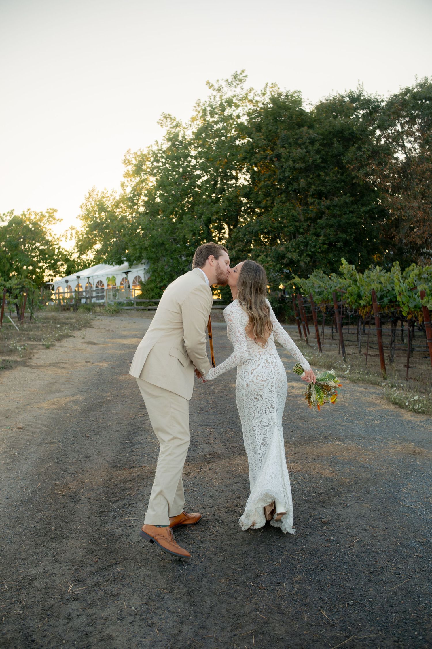 Bride and groom sharing a kiss on a vineyard path at golden hour with lush greenery behind them.