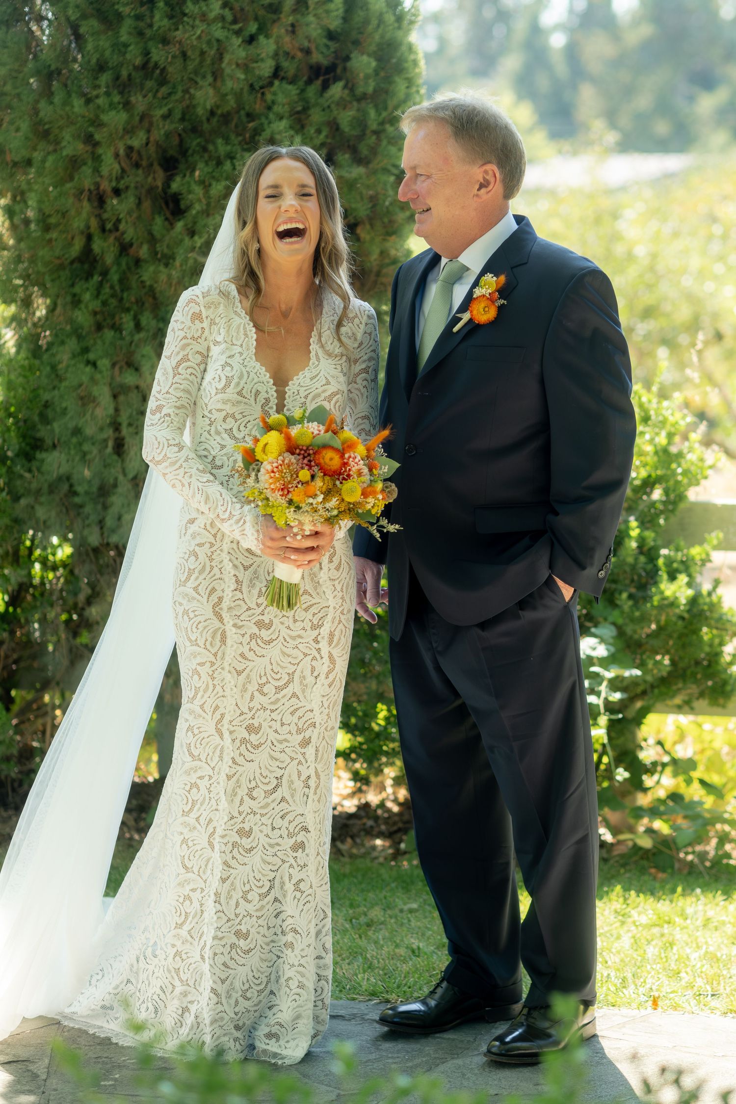 Bride in lace gown laughing with older man in dark suit holding autumn bouquet outdoors.