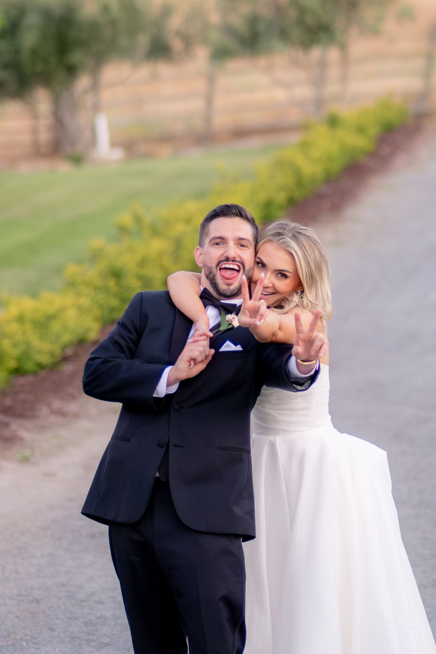 Joyful bride and groom show off wedding rings while laughing on a scenic vineyard pathway.