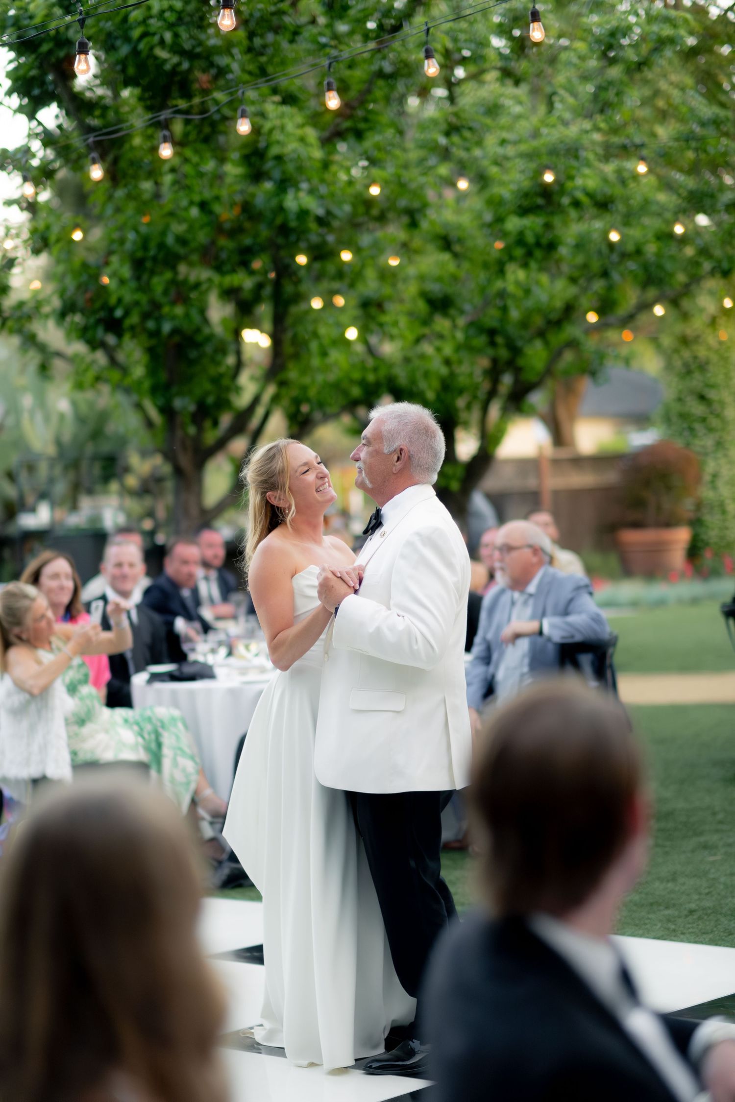 Bride dancing with older gentleman in white tuxedo jacket on dance floor at outdoor evening reception.