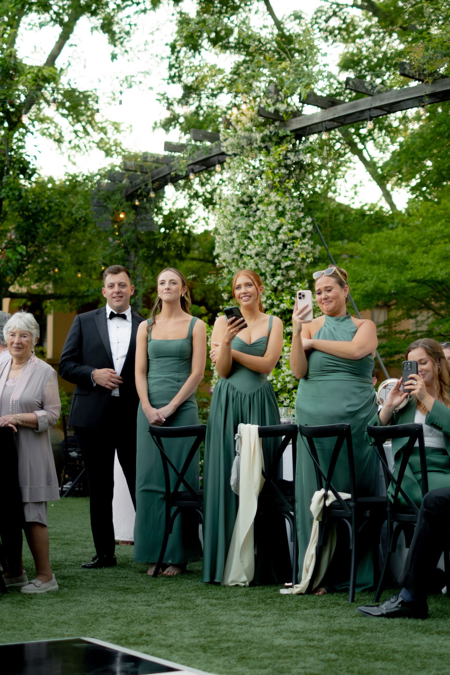 Bridesmaids in elegant green satin gowns stand with groomsman watching wedding ceremony in lush garden venue.