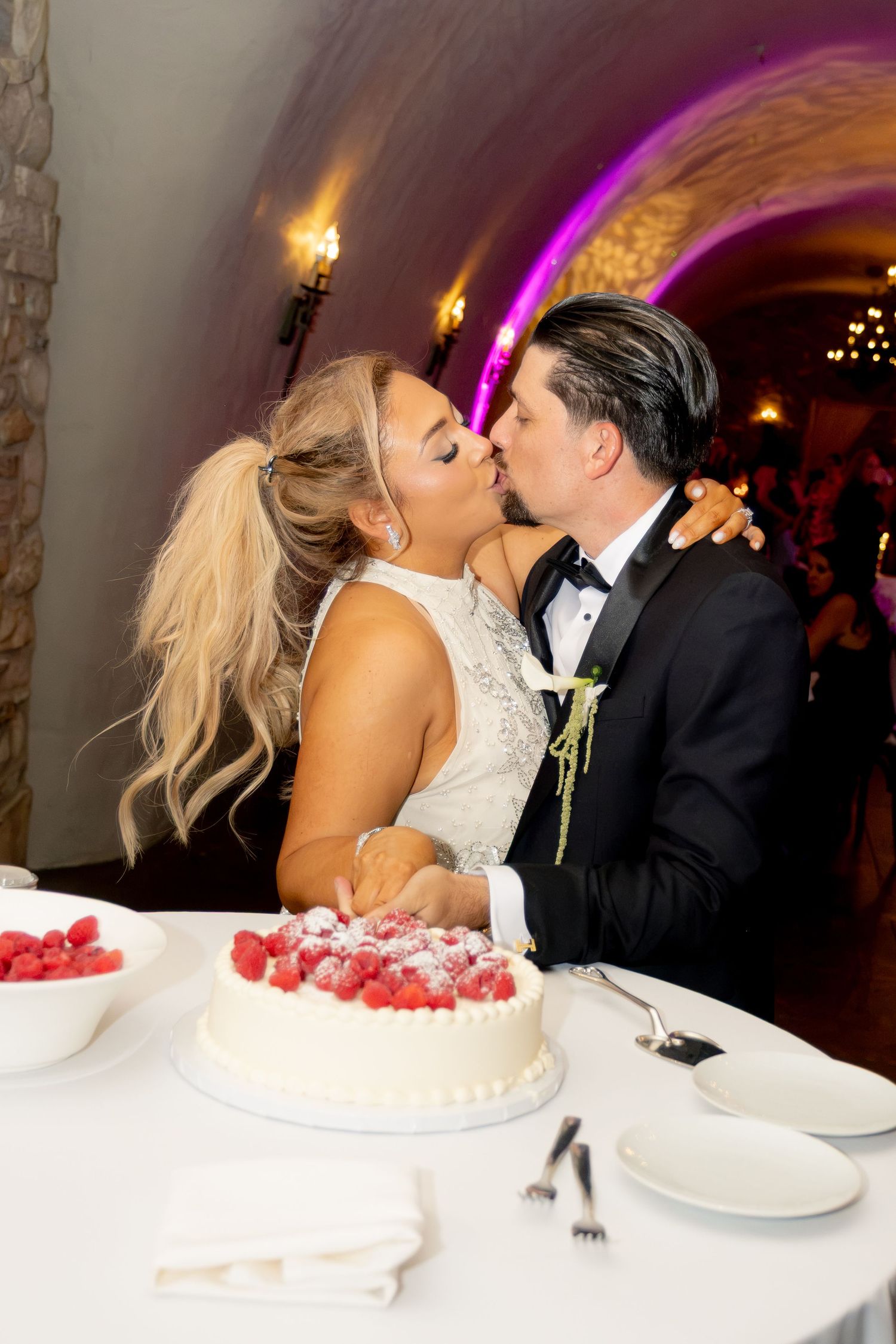 Bride and groom share romantic kiss over white raspberry wedding cake in arched stone reception venue.