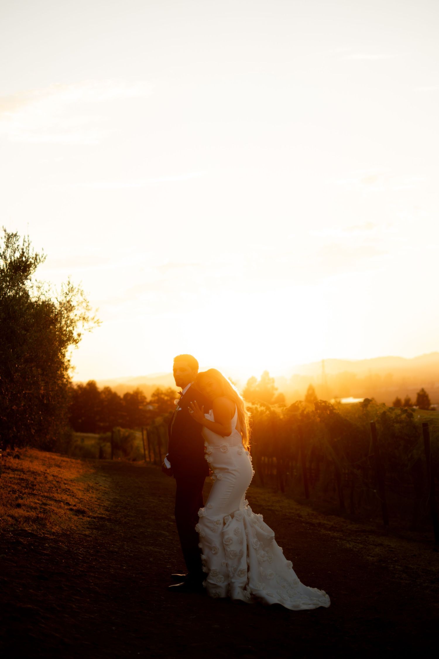 Bride and groom embrace at golden sunset overlooking a scenic vineyard landscape on their wedding day.