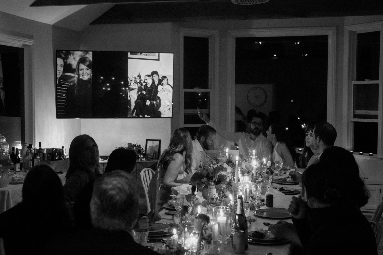 Wedding reception dinner in candlelit room with guests seated at table and photos projected on wall.