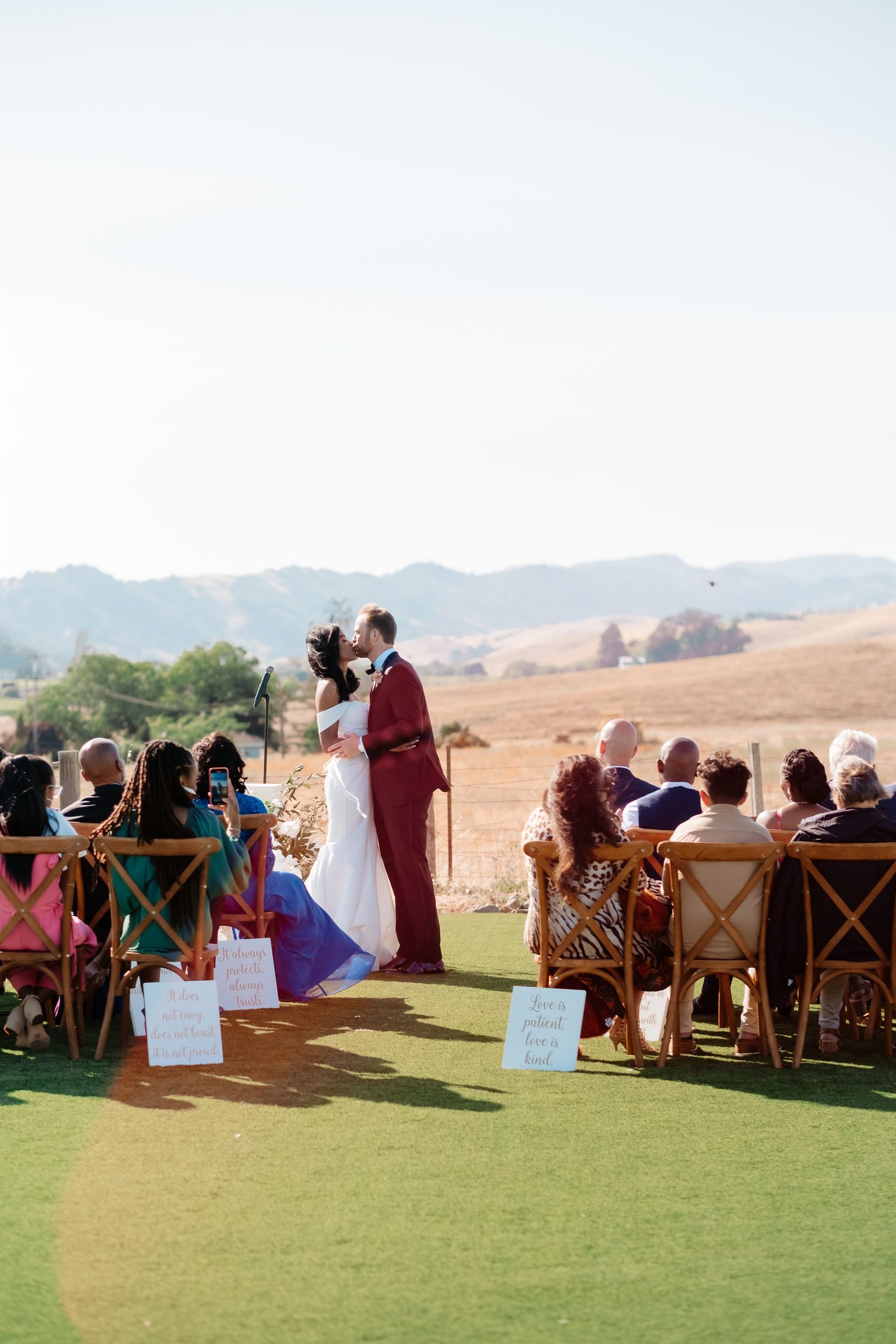 Bride and groom share first kiss at outdoor wedding ceremony with golden hills backdrop.