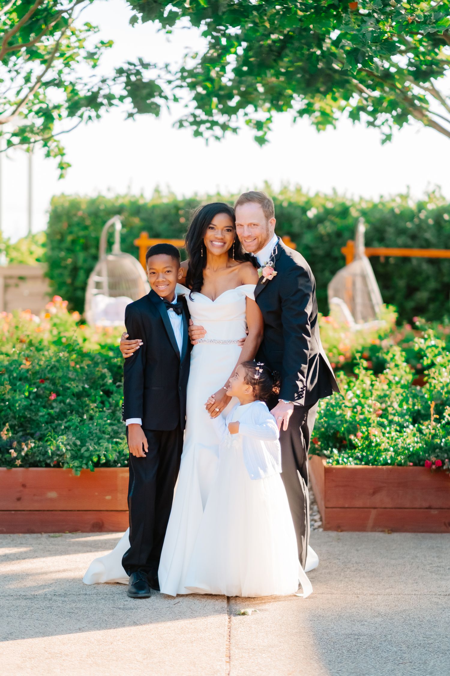 Interracial wedding couple poses outdoors with young boy in tuxedo and flower girl in white dress.