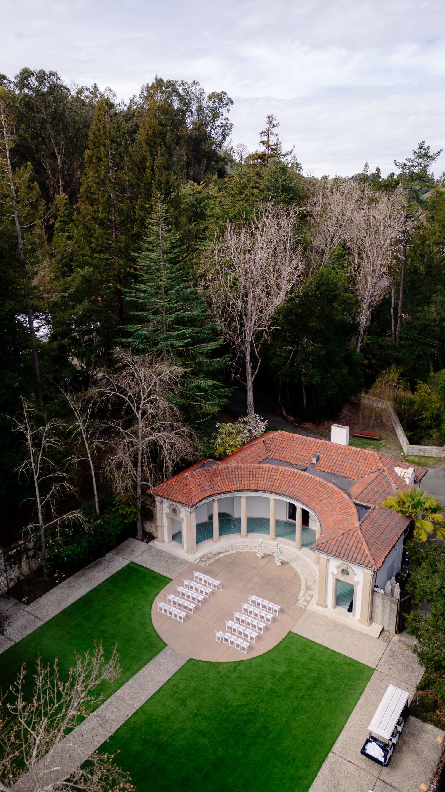 Aerial view of elegant Mediterranean-style event venue with red tile roof, white chairs arranged outdoors, surrounded by lush green trees.