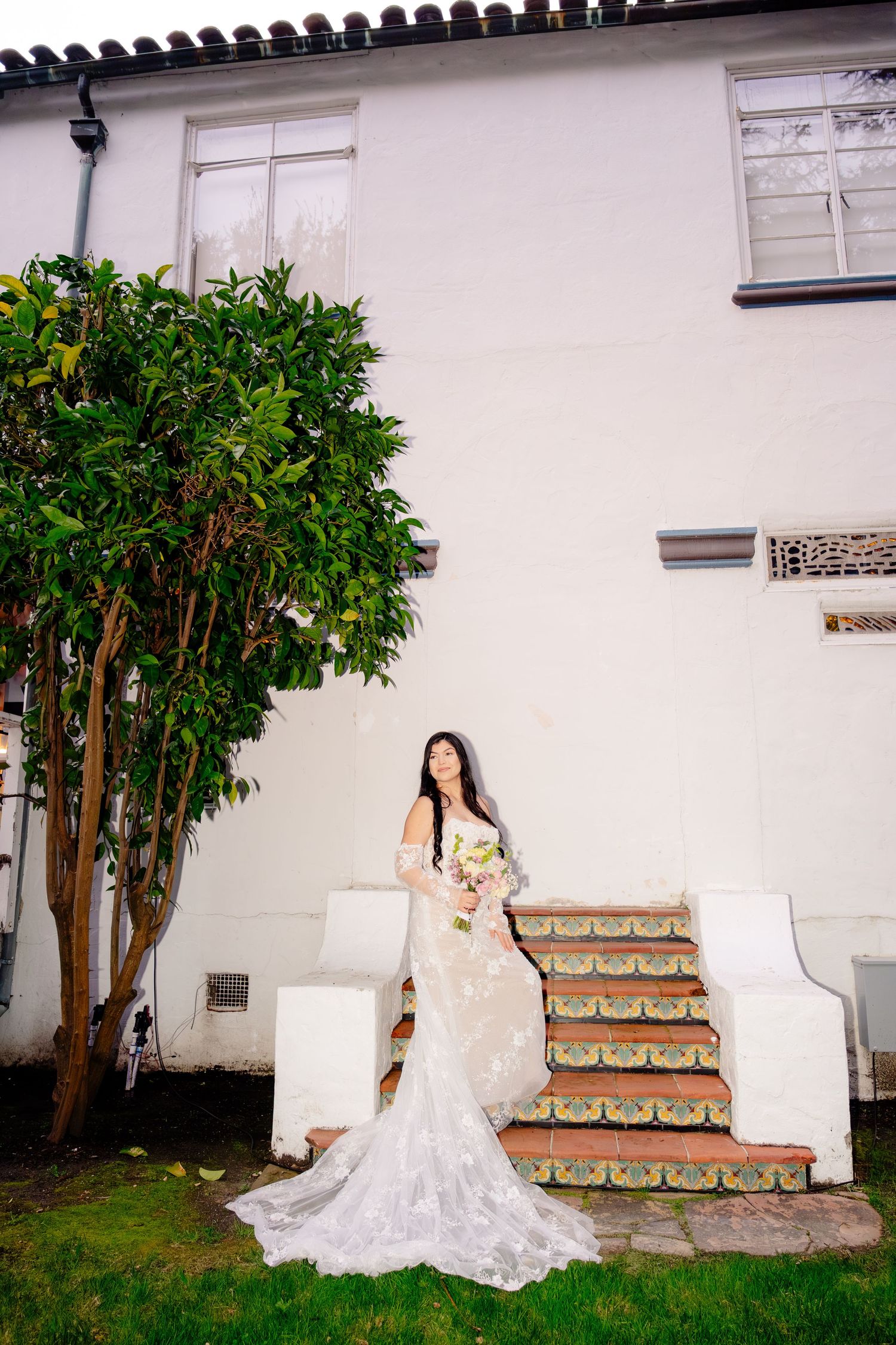Bride in long lace mermaid wedding gown poses with bouquet beside steps and white building.