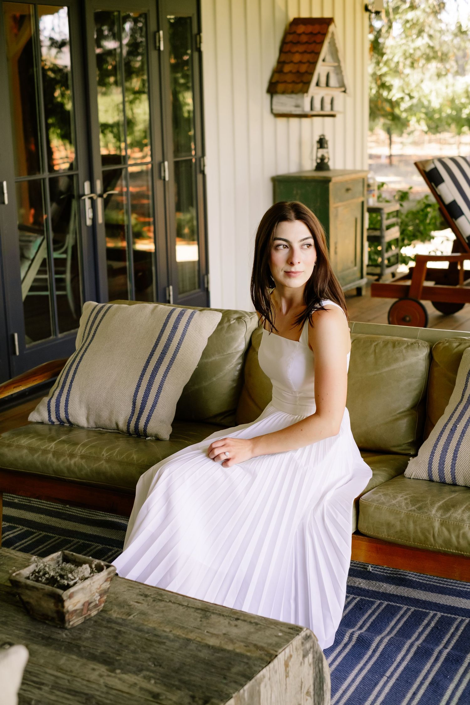 Young woman in white pleated dress sitting on olive green sofa on rustic porch with striped pillows.