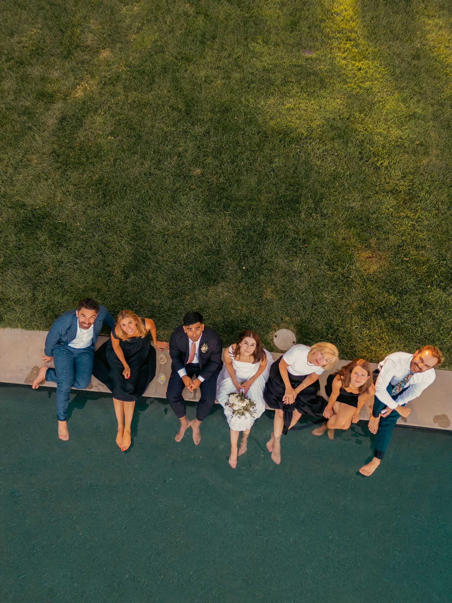 Aerial view of a wedding party seated along a pathway edge surrounded by green grass, smiling upward.