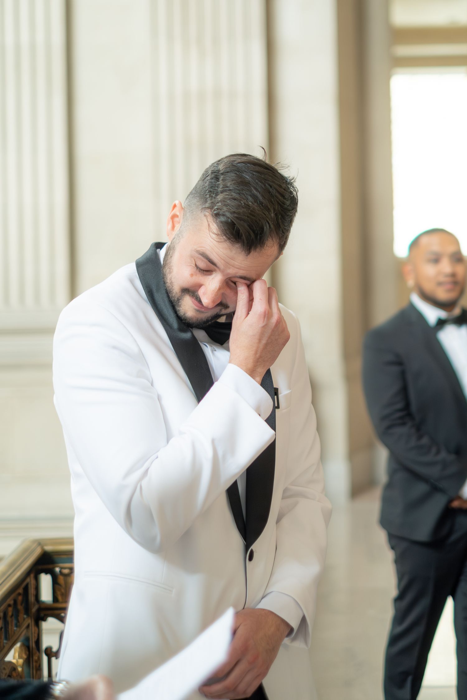 Emotional groom in white tuxedo wiping tears while reading vows at elegant wedding ceremony.