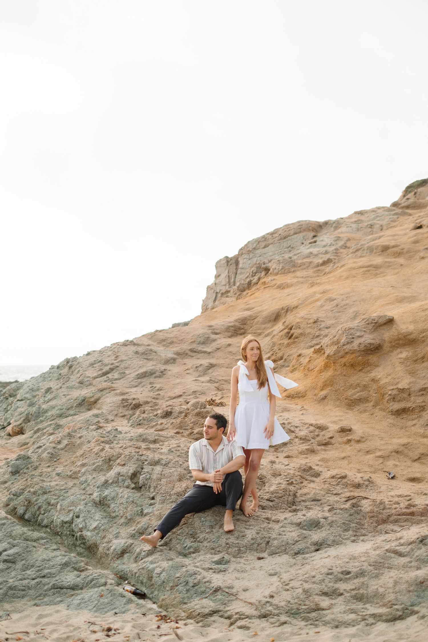 Couple posing on sandy coastal rock formations with dramatic cliffs and ocean scenery behind them.