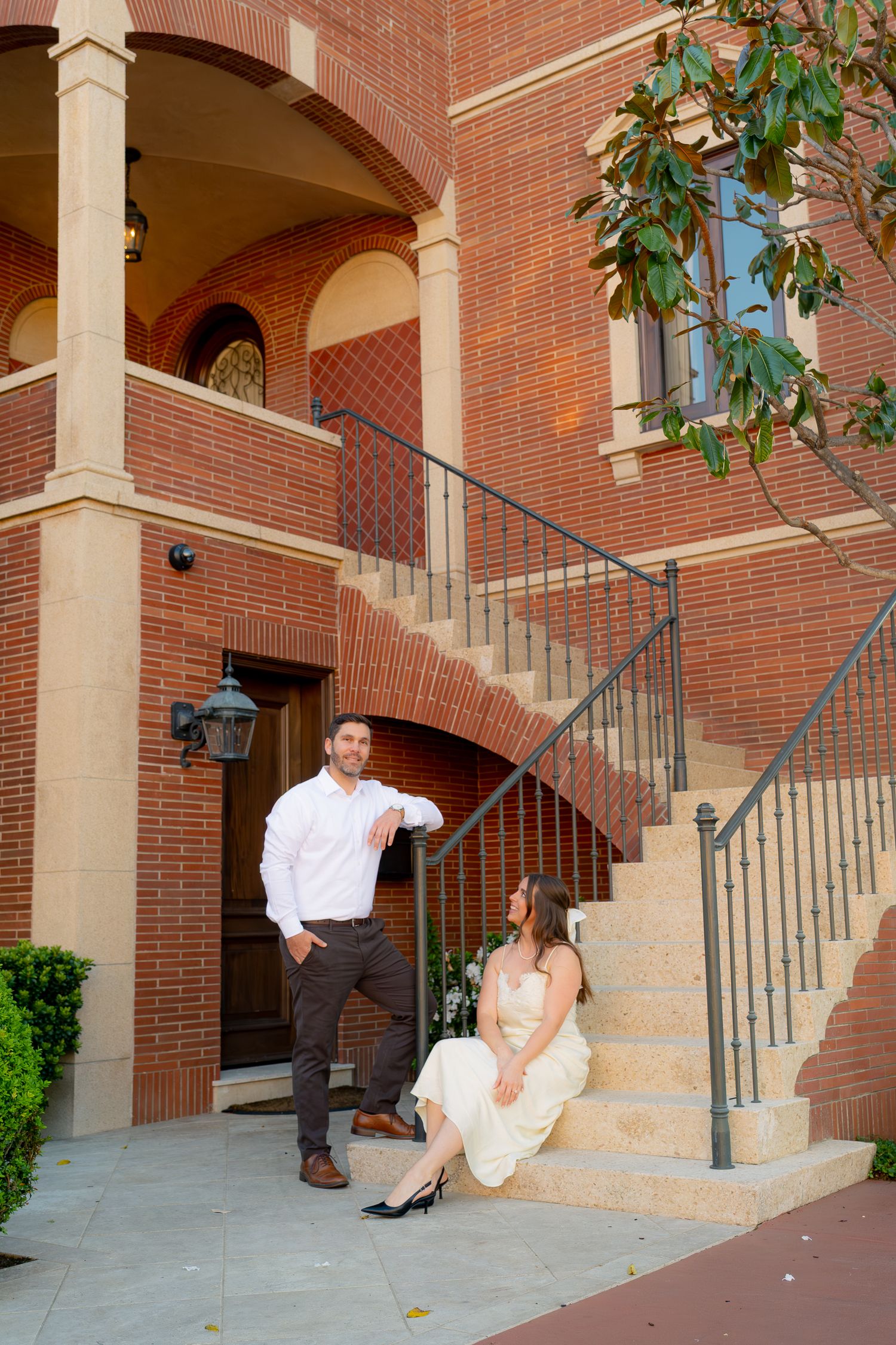 Couple posing on brick building steps, man in white shirt standing while woman in cream dress sits on stairs.