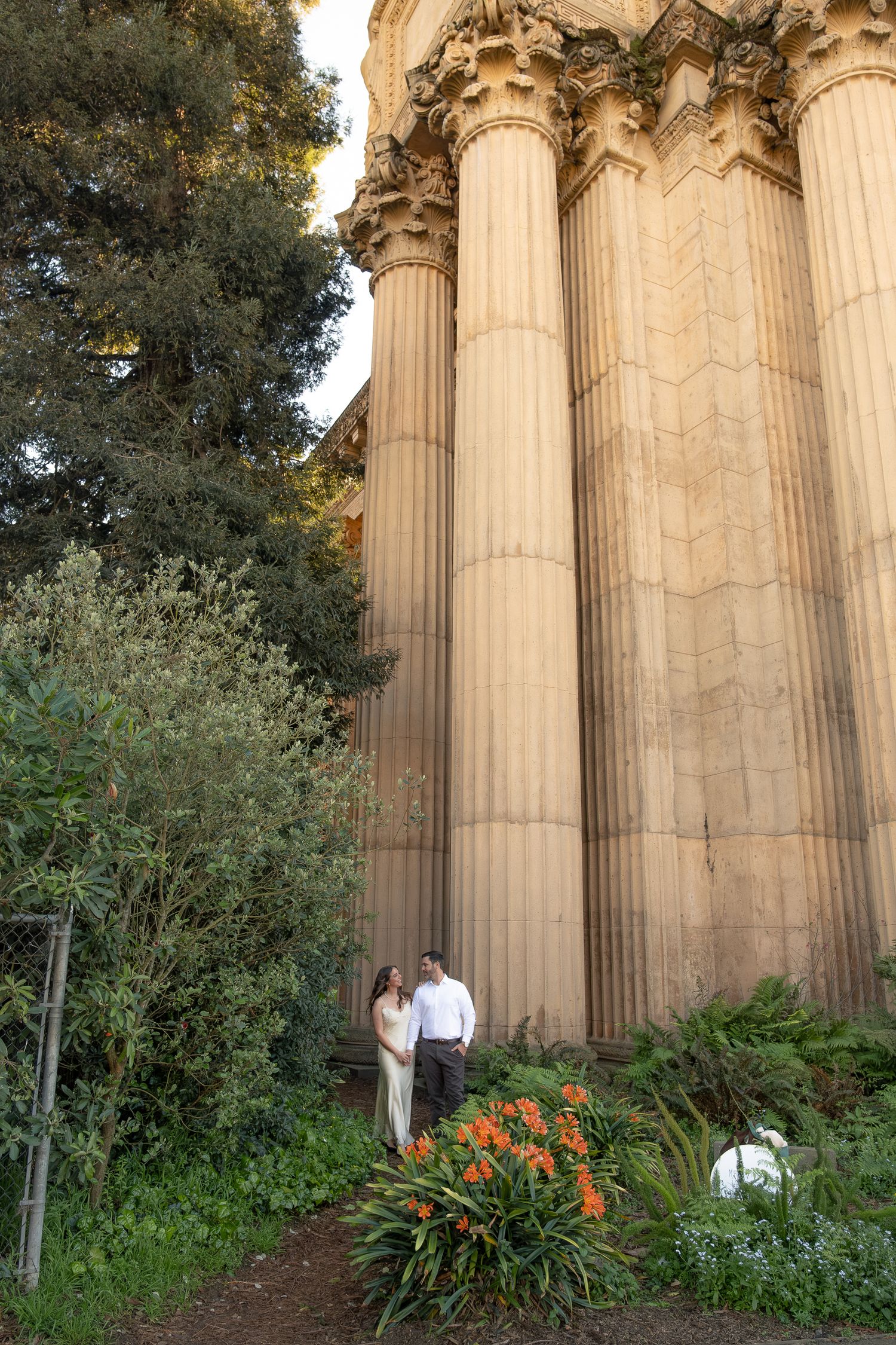 Couple standing among lush greenery beside towering ornate columns at the Palace of Fine Arts in San Francisco.