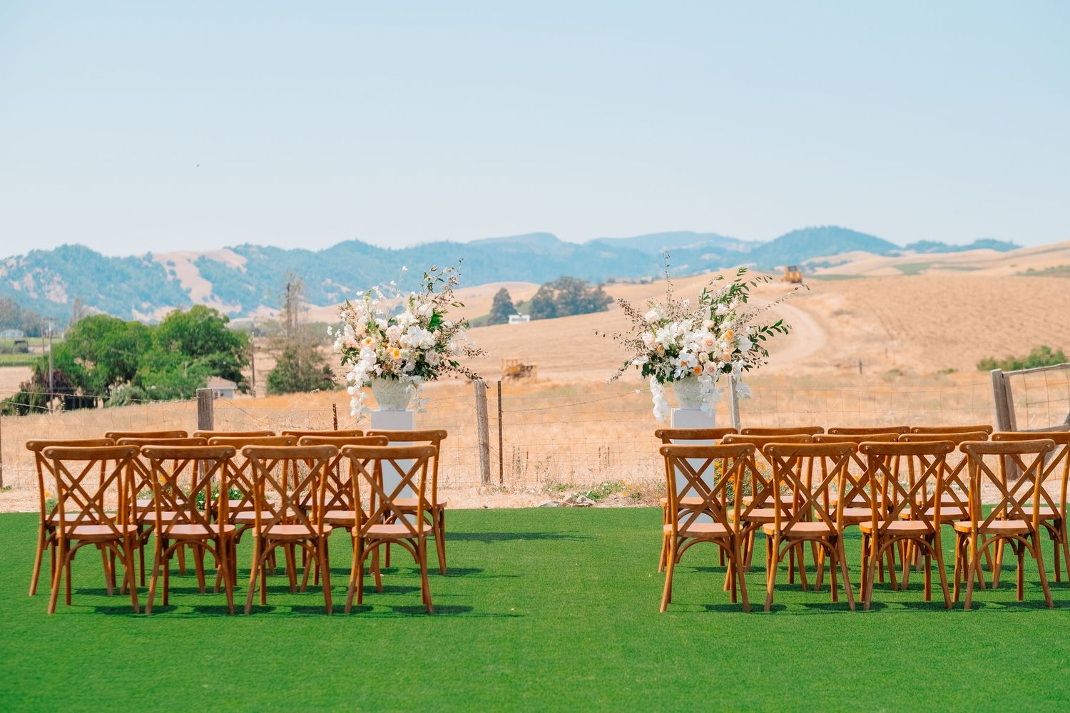 Outdoor wedding ceremony setup with wooden chairs and floral arrangements on green lawn with golden hills backdrop.