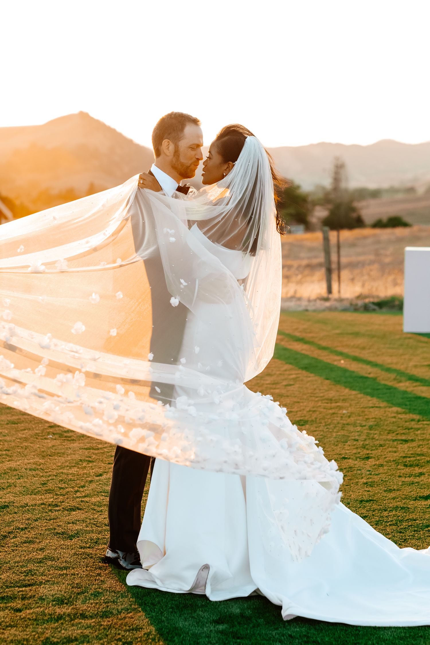 Bride and groom kiss at golden sunset with floral veil billowing in the breeze outdoors.