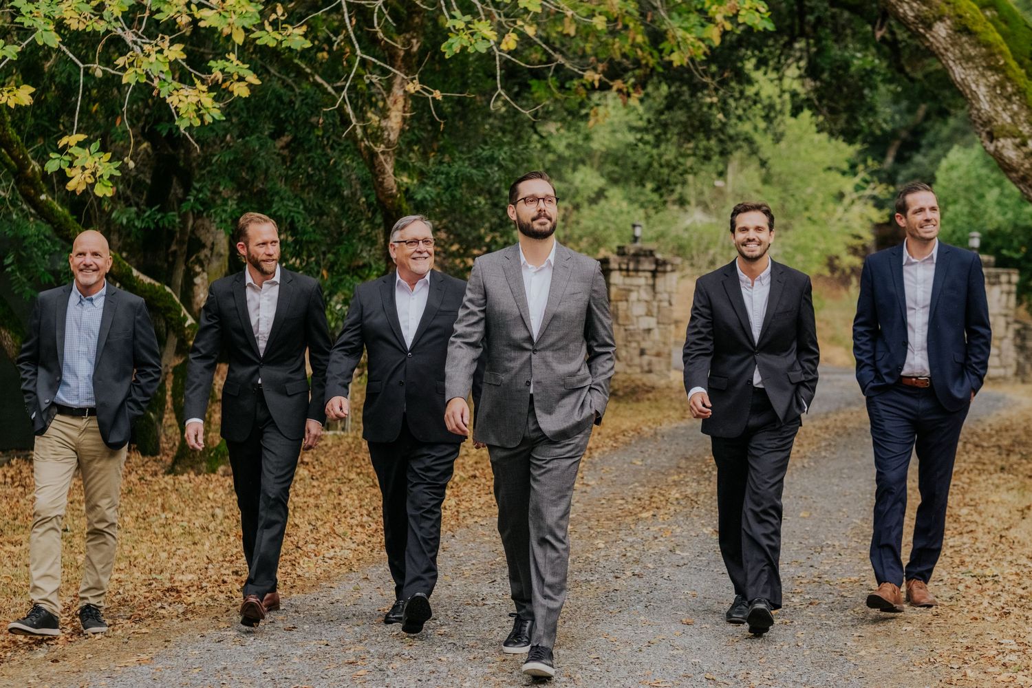 Seven groomsmen in dark suits walking together outdoors on a gravel path surrounded by lush green trees.