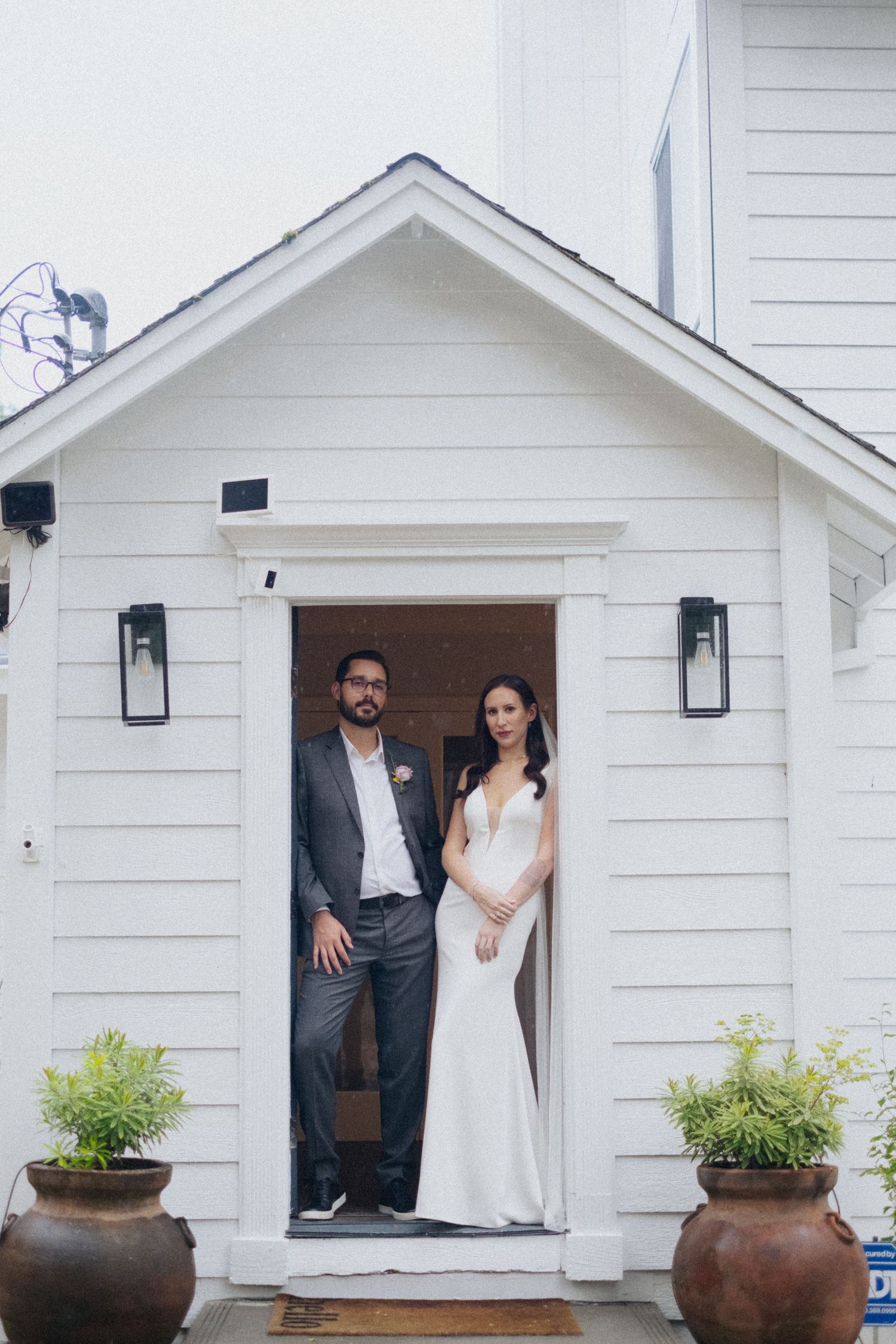 Bride and groom posing in doorway of white clapboard building with potted plants on wedding day.