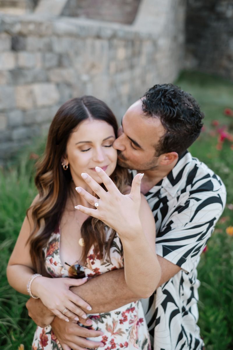 Couple celebrating engagement outdoors, woman showing diamond ring while man kisses her cheek near stone wall.