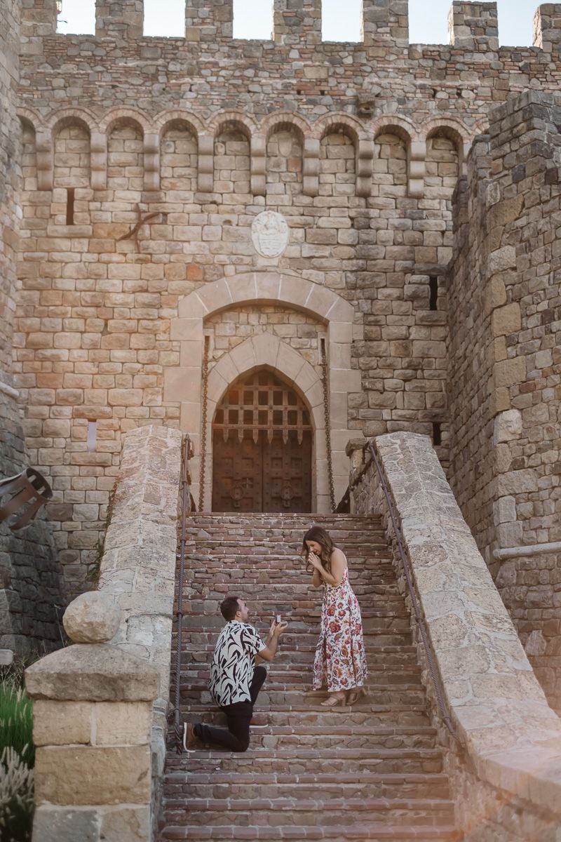 Man proposing on one knee to woman in floral dress on castle stone staircase.