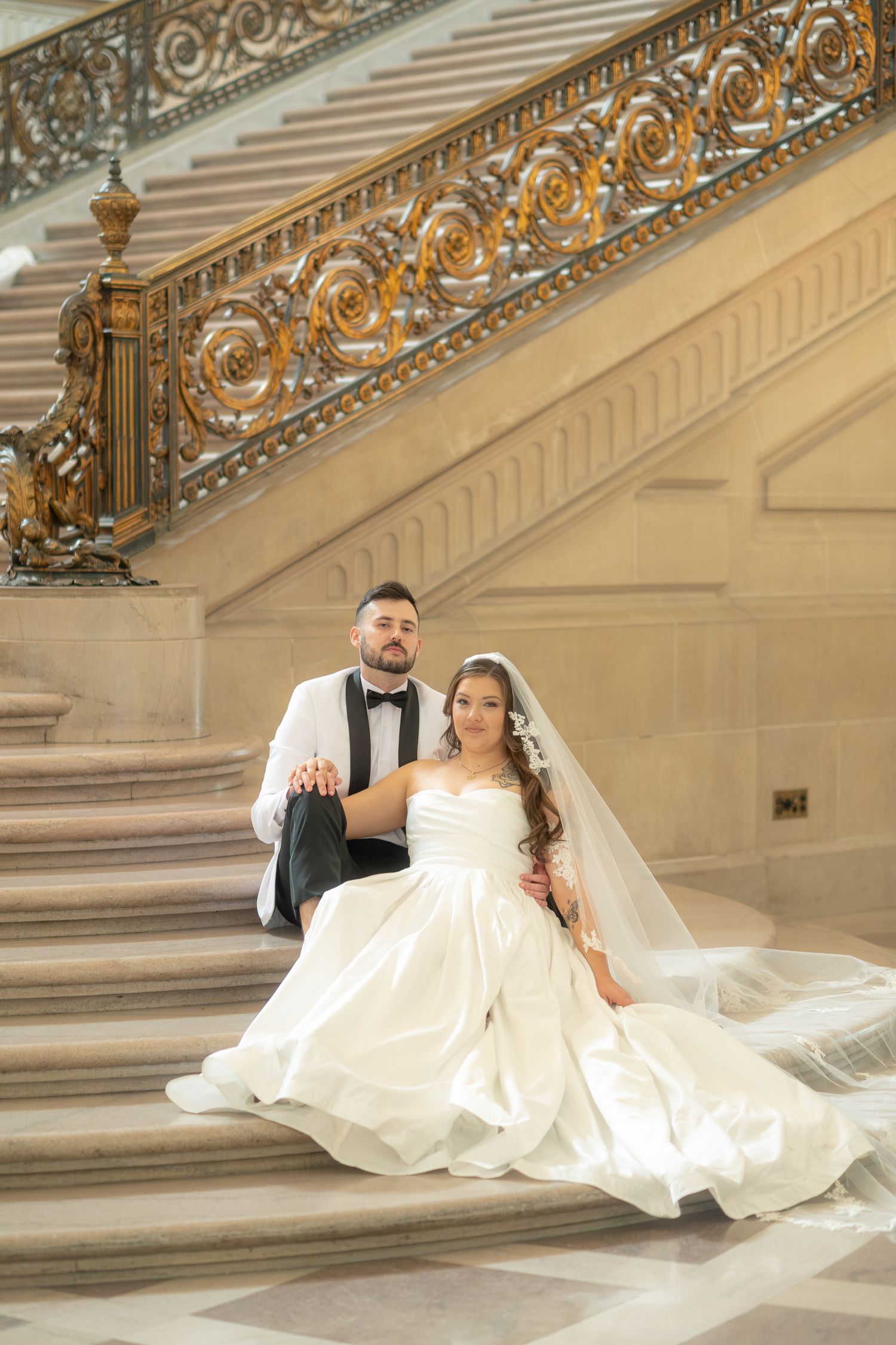 Bride and groom sitting on ornate golden staircase in elegant wedding attire at San Francisco City Hall.