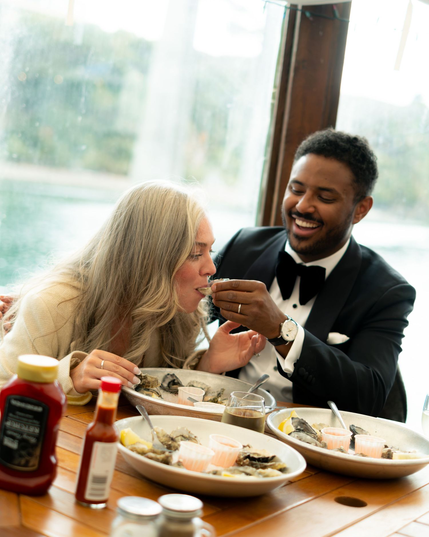 Groom in tuxedo feeds bride oysters at a waterfront restaurant during their wedding celebration.