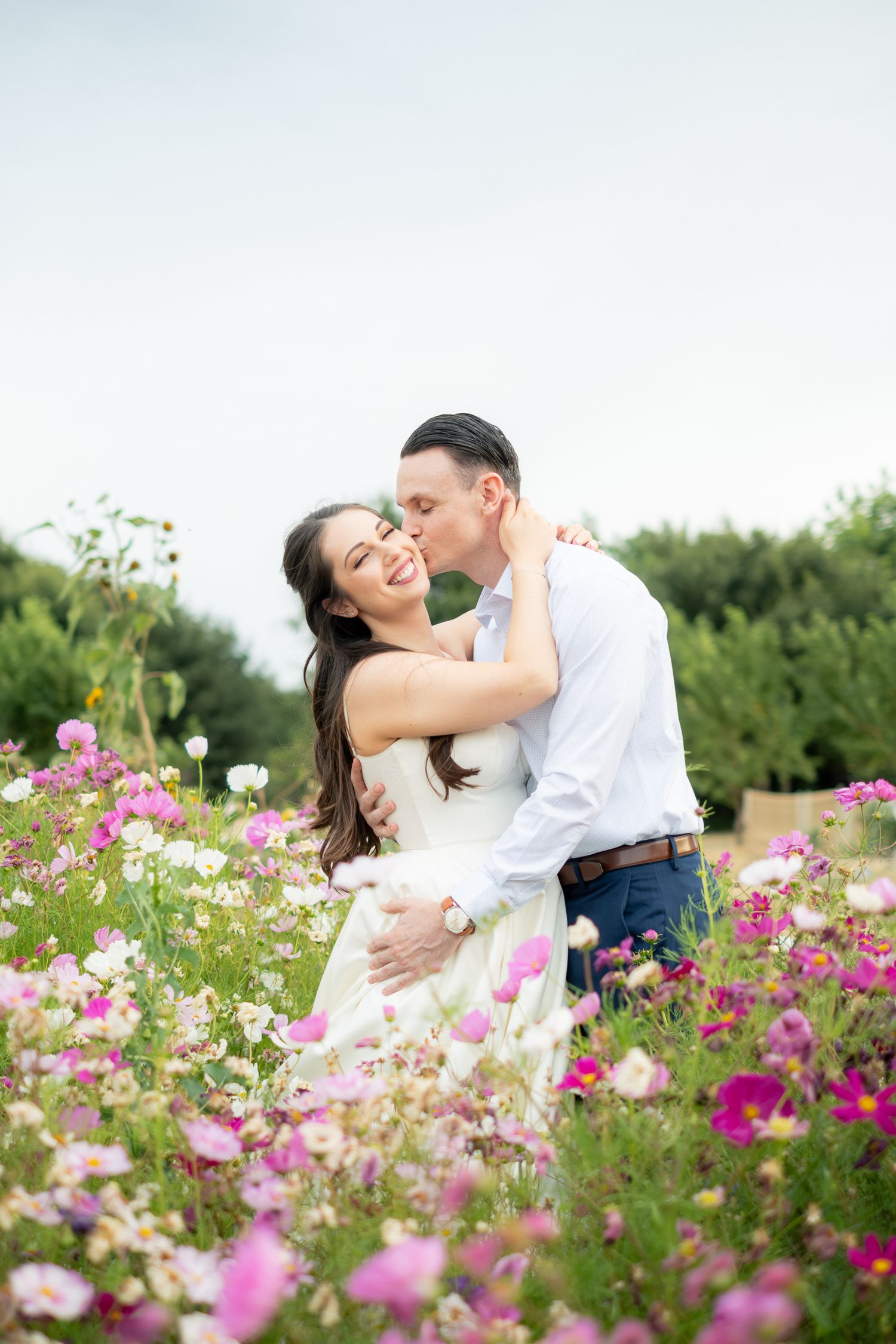 Couple embracing in a field of pink and white cosmos flowers, groom kissing bride on cheek as she smiles joyfully.