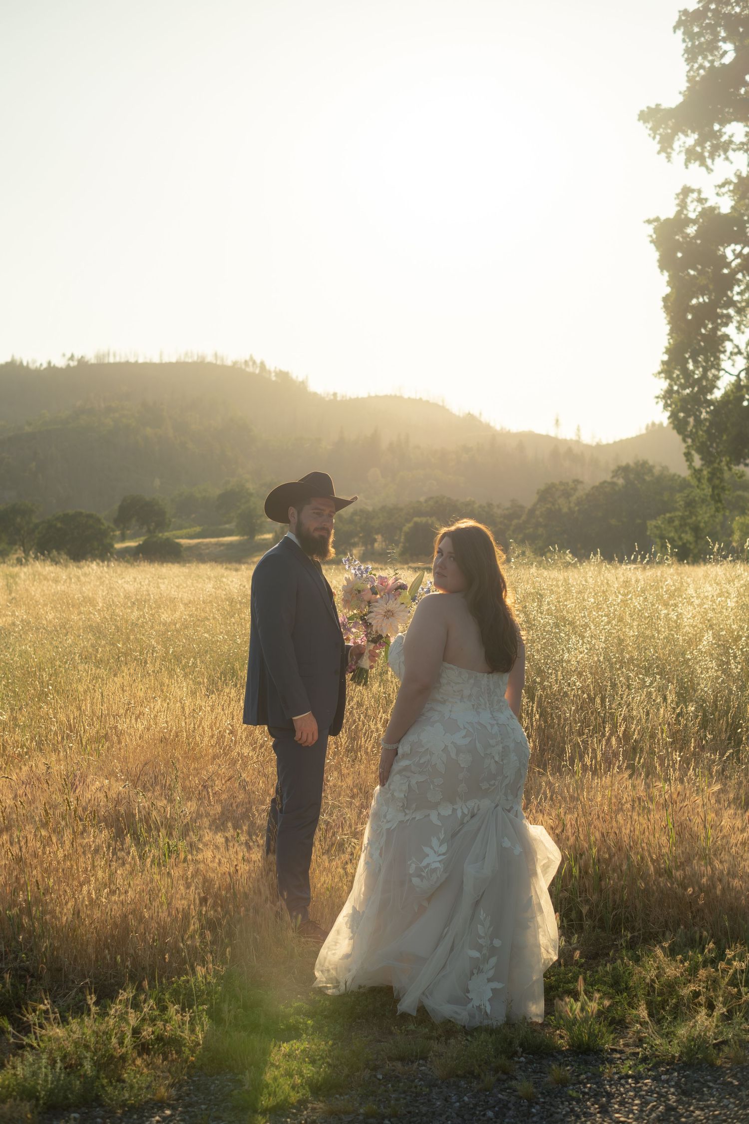 Bride and groom stand in golden field at sunset with mountain views behind.