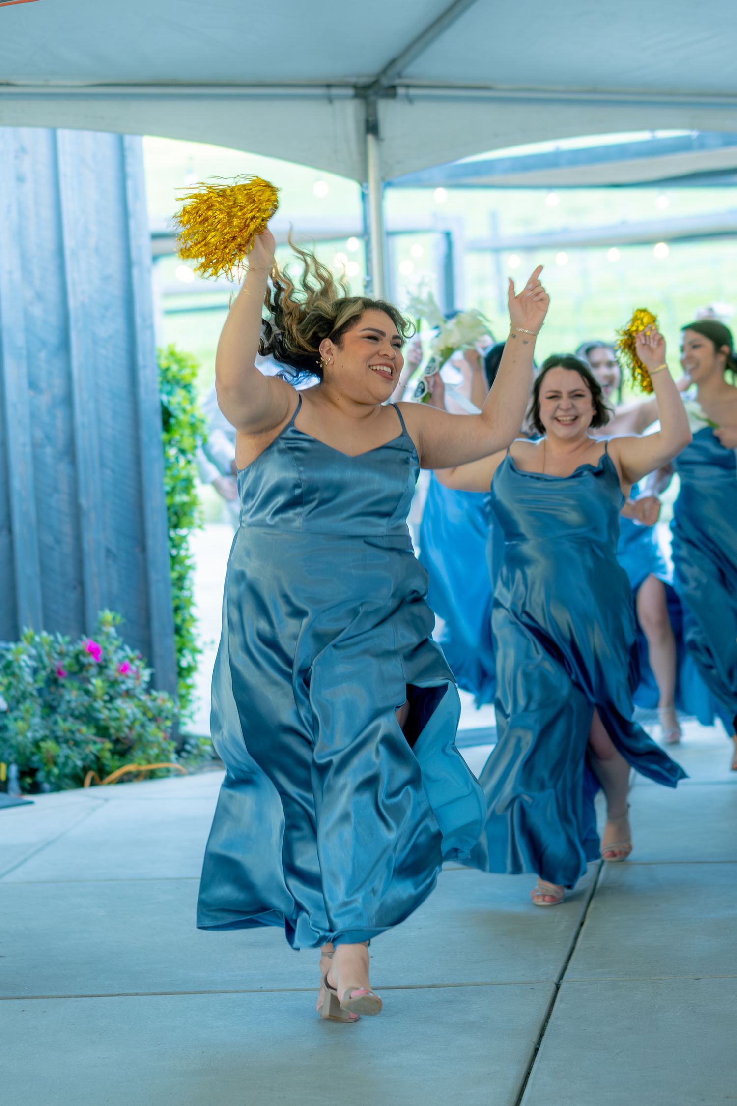 Bridesmaids in blue satin dresses dance energetically with gold pom-poms at wedding reception.