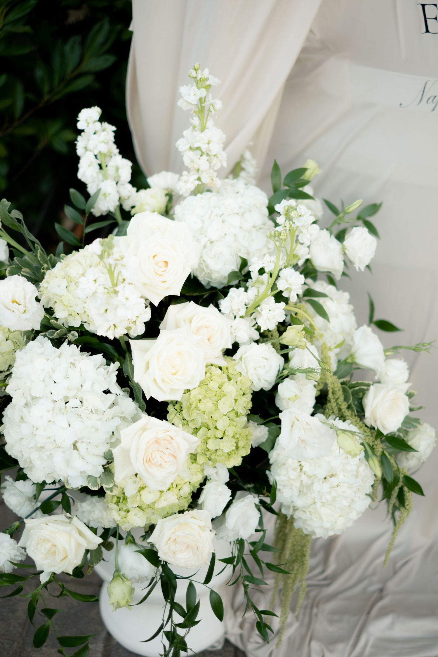 Lush white wedding floral arrangement featuring roses, hydrangeas, and greenery in white vase.
