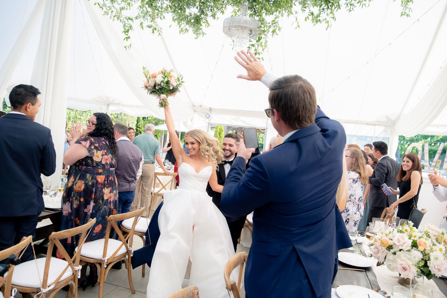 Bride raises bouquet triumphantly as she enters tented wedding reception to cheering guests.