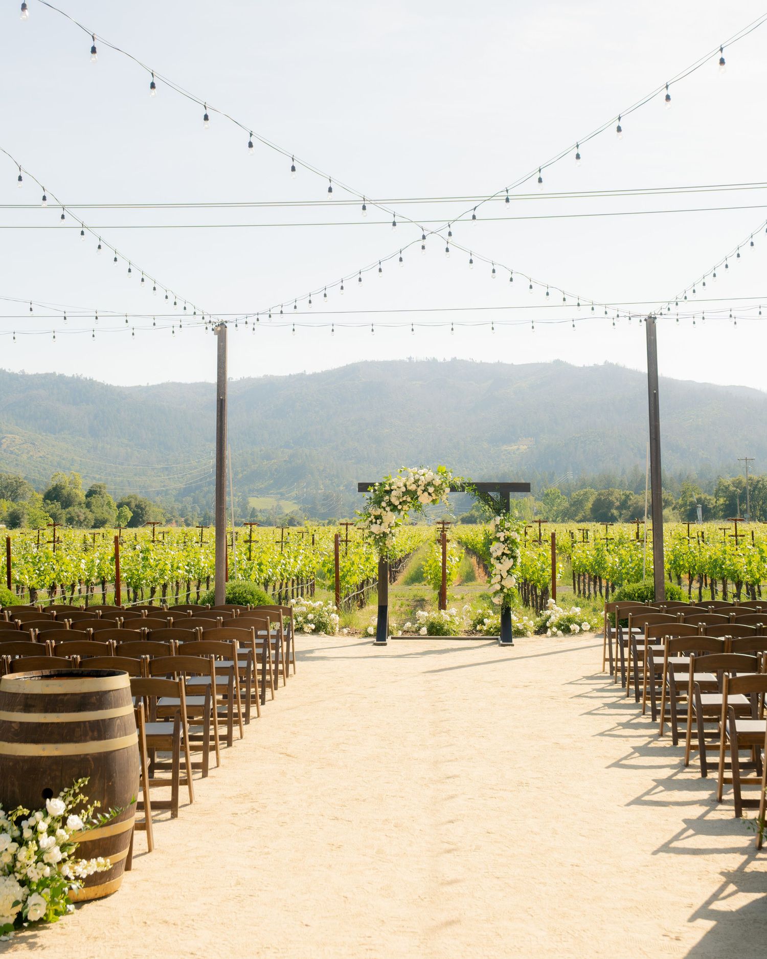 Outdoor vineyard wedding ceremony setup with wooden barrel aisle markers, string lights and floral arch.