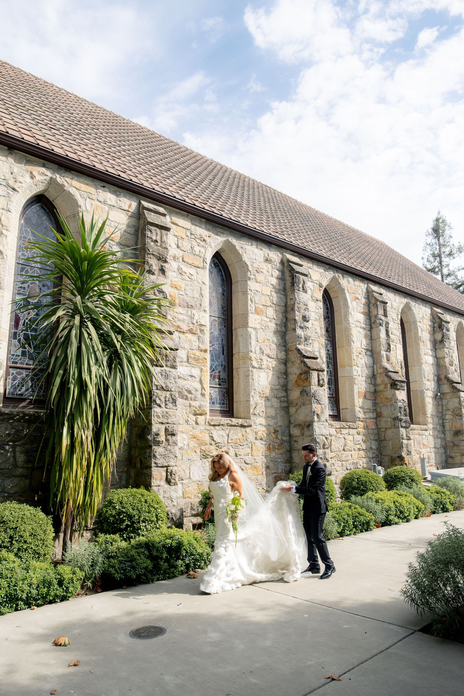Bride and groom share a romantic moment outside a historic stone church with arched windows and lush greenery.