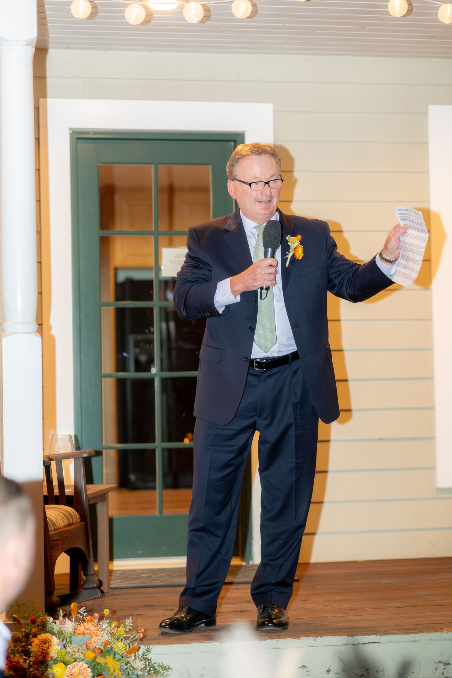 Man in dark suit giving a speech at wedding reception holding microphone and notes with yellow boutonniere.