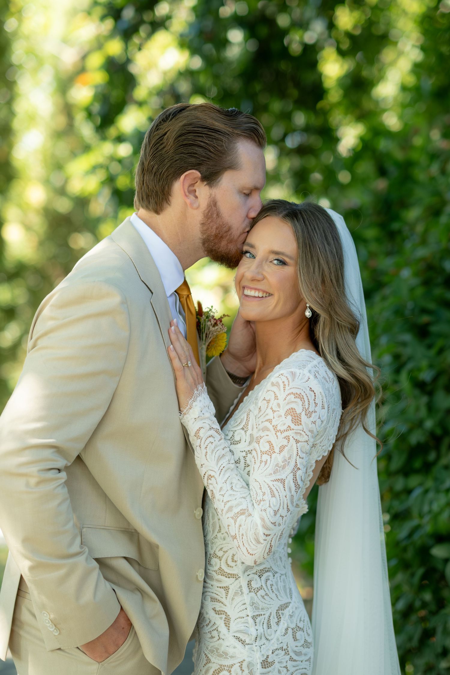 Groom in tan suit kissing bride on cheek as she smiles in lace wedding dress before green foliage.