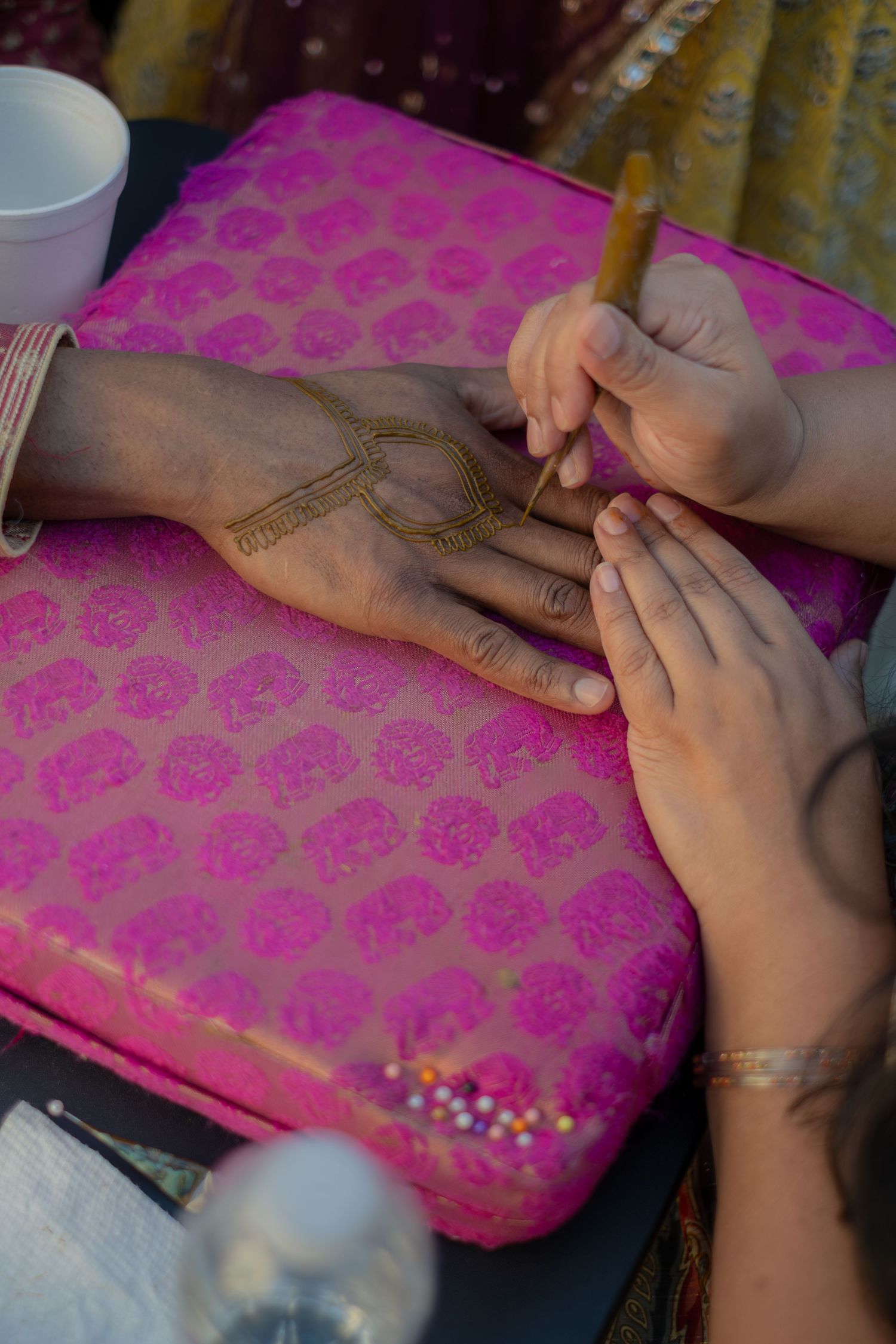 Artist applying intricate henna mehndi design on a hand resting on a pink embroidered cushion.