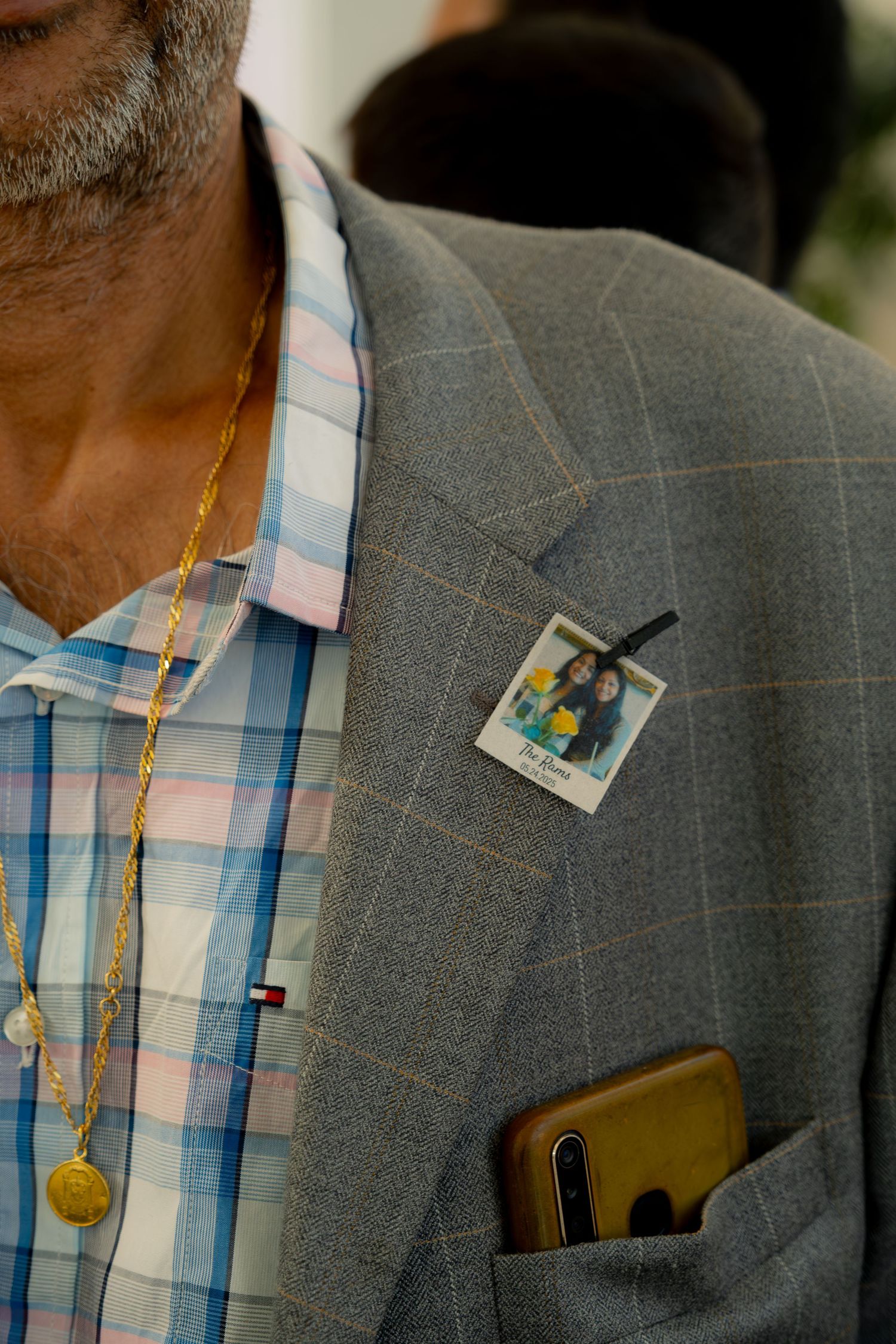 Close-up of a man in grey plaid blazer wearing a polaroid photo pin and gold chain necklace at a wedding.
