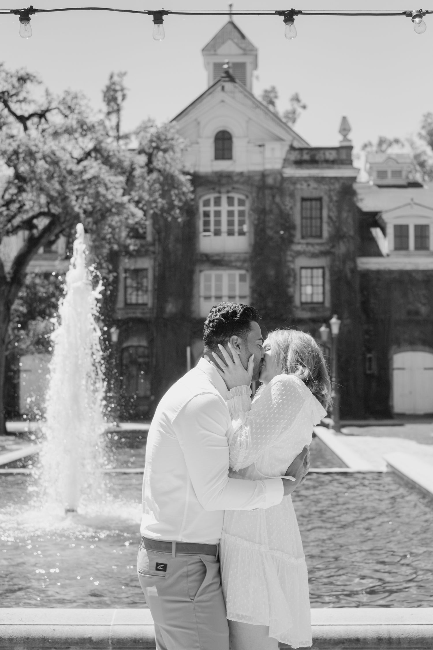 Black and white photo of couple kissing near a fountain with a historic building in the background.