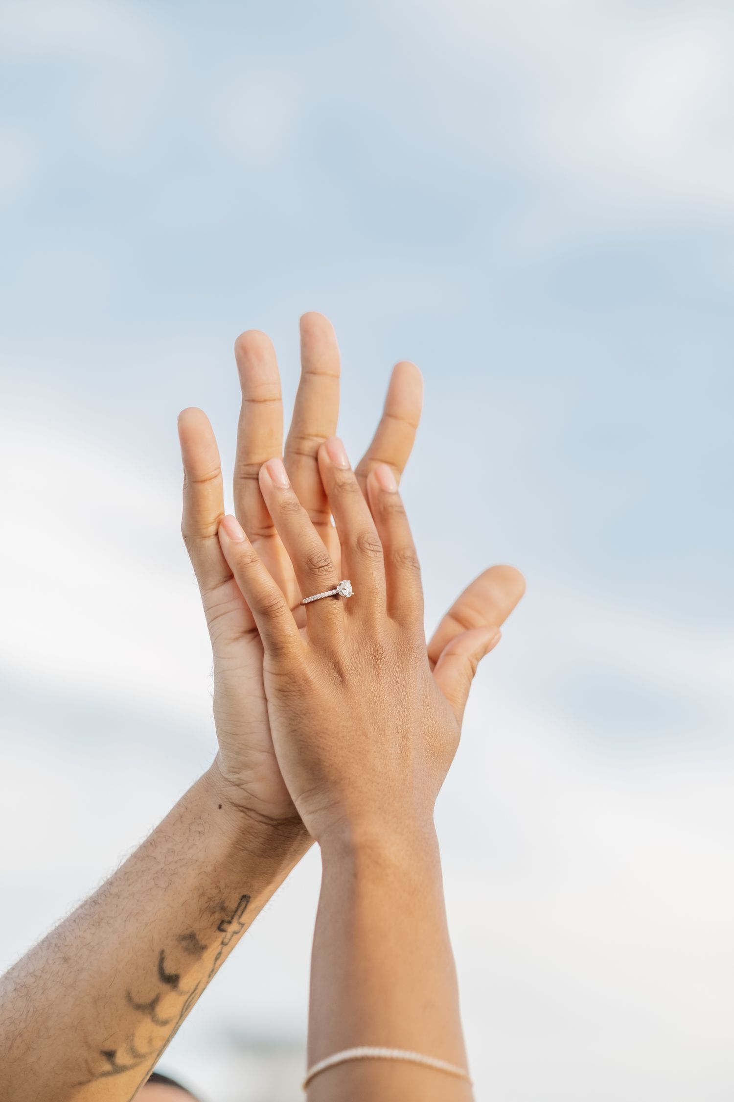 A couple raises their intertwined hands toward a cloudy sky, showcasing an engagement ring and tattoo.