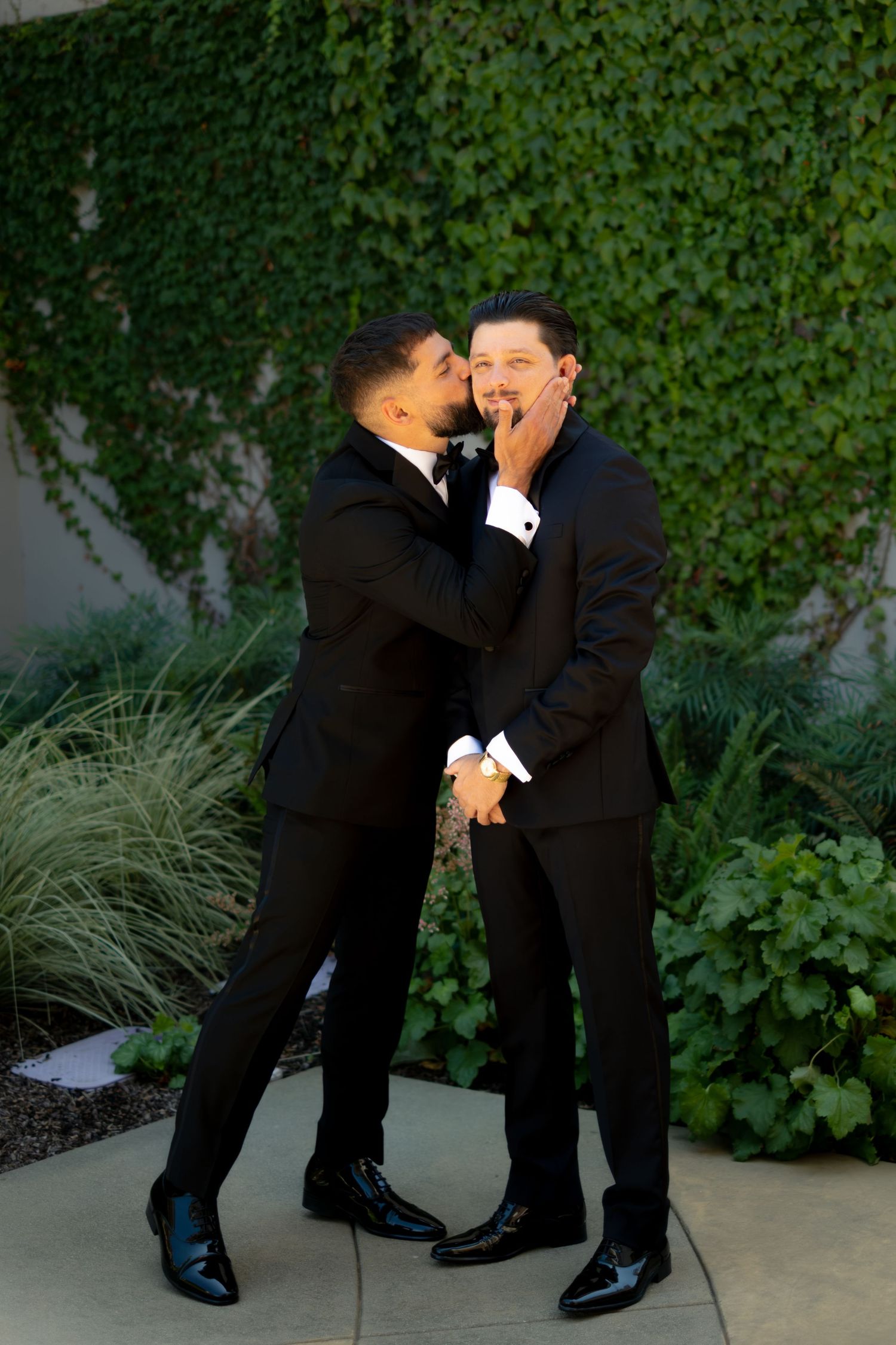 Two grooms in black tuxedos share a tender romantic moment in front of a lush ivy-covered wall.