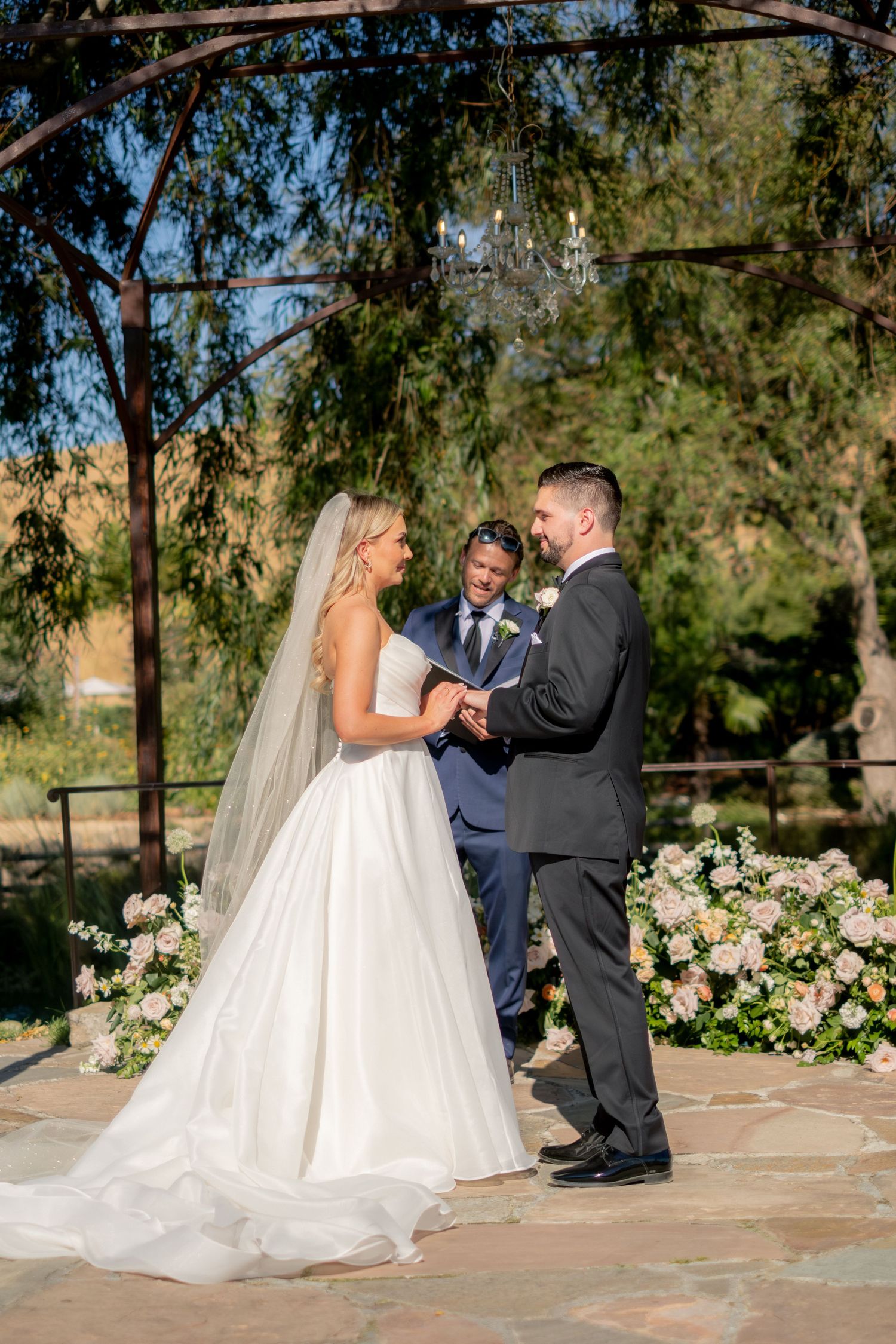 Bride and groom exchange vows under a floral arbor with chandelier at an outdoor wedding ceremony.