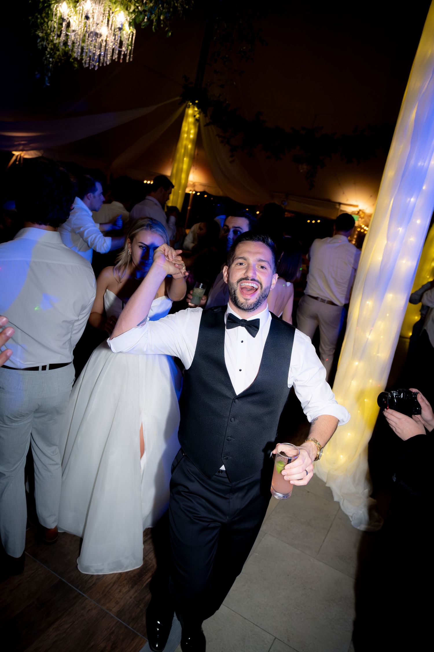 Groom dancing joyfully with guests under string lights at an energetic wedding reception party.