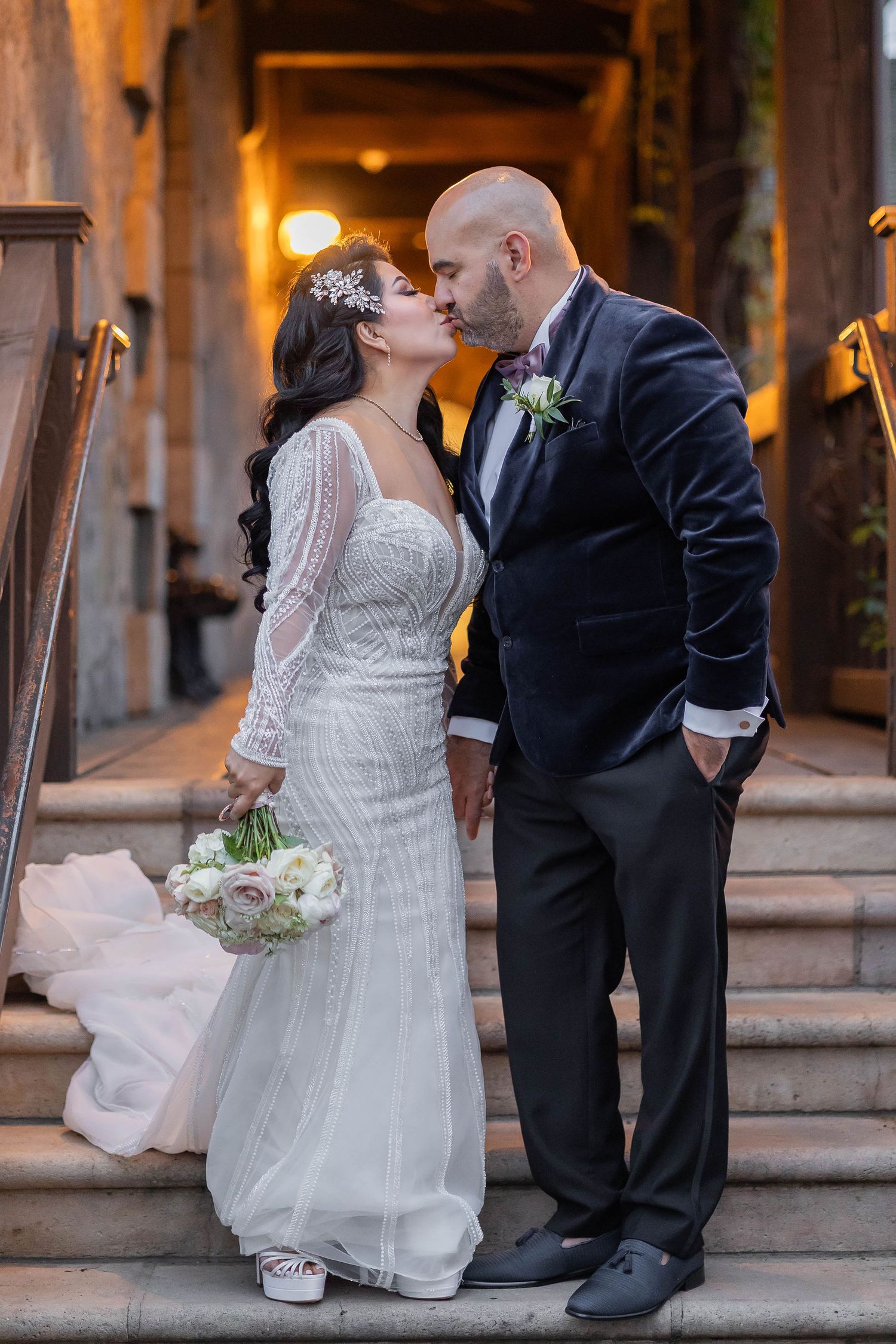Bride in lace long-sleeve gown and groom in navy suit sharing kiss on stone staircase.