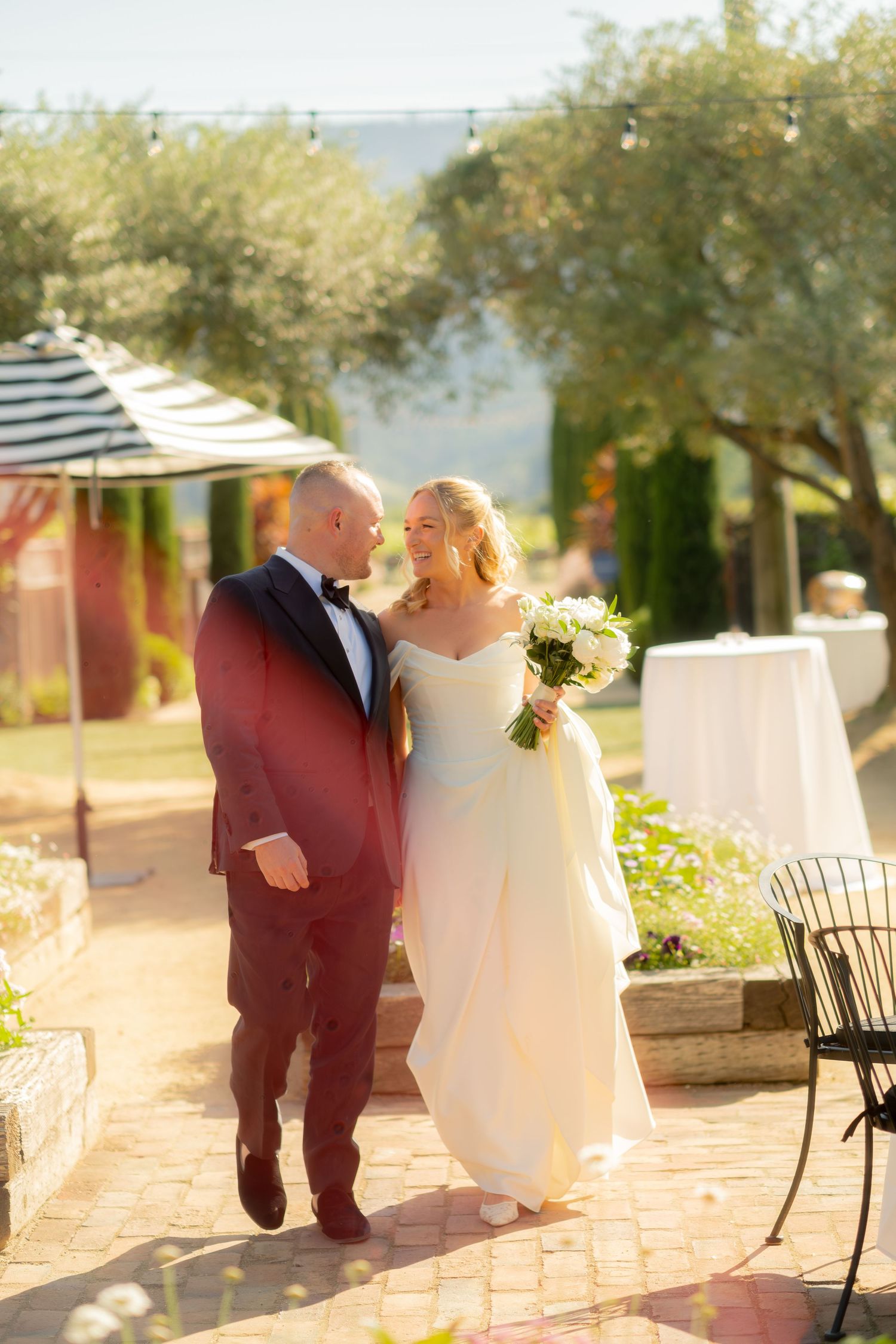 Bride and groom walking together outdoors at a sunny vineyard wedding venue, bride holding white bouquet.