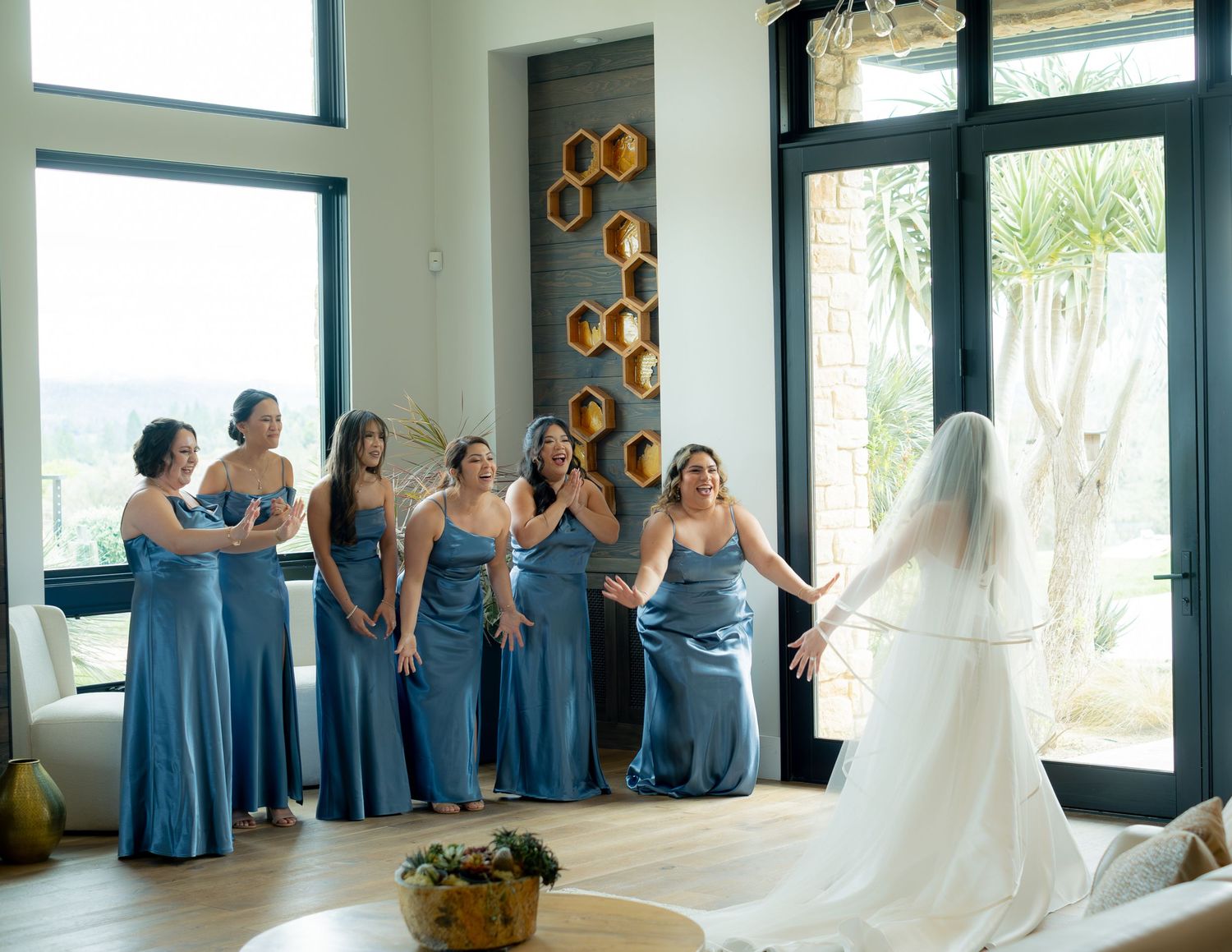 Bridesmaids in blue dresses react with joy as bride in white gown and veil reveals herself.